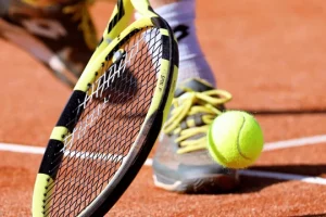 Les activités et loisirs autour de Lacanau. Deux pieds d'un joueur de tennis sur un terrain de terre battue, il reprend en demie volée une balle de tennis jaune avec une raquette noire et jaune.