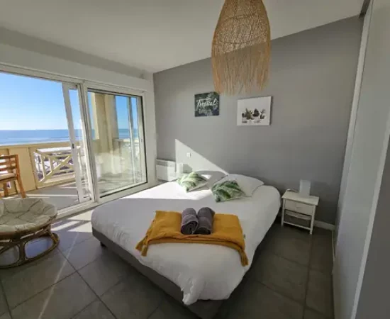 Appart Hôtel Pasino N°4, chambre appartement n°4 donnant sur la terrasse vue mer, 2 chevets, 2 lampes de chevet, 2 coussins décoration sur le lit, un plaid jaune, 2 serviettes posées sur le lit. Décorations au mur et plafond. sur la gauche un fauteuil en rotin. A l'arrière de la photo vue sur la terrasse et l'océan