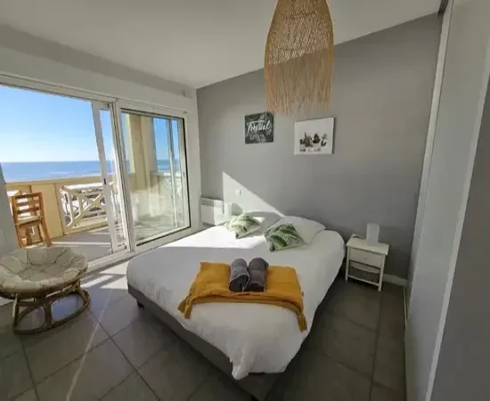 Appart Hôtel Pasino N°4, chambre appartement donnant sur la terrasse vue mer, 2 chevets, 2 lampes de chevet, 2 coussins décoration sur le lit, un plaid jaune, 2 serviettes posées sur le lit. Décorations au mur et plafond. sur la gauche un fauteuil en rotin. A l'arrière de la photo vue sur la terrasse et l'océan