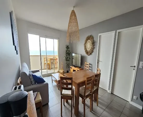 Appart Hôtel Pasino N°2, pièce à vivre de l'appartement numéro 2, elle est lumineuse avec ses murs blancs, elle est meublée avec des meubles en teck: une table, ses six chaises et leurs coussin d'assise beiges. A l'arrière la partie salon: sa table de salon en teck , son meuble TV et la TV grand écran. des éléments de décoration au mur, un palmier au fond de la pièce. une baie vitrée donne sur la terrasse vue mer et ses meubles en teck